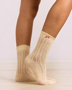 Chaussettes Merci Paris Damson Madder en maille pointelle écru avec broderie DM rouge, portées sur les pieds